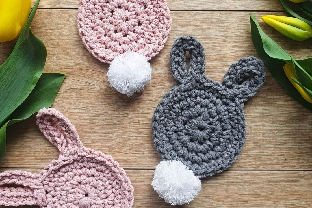 Drei hasenfärmige Untersetzer aus Makrameegarn in Rosa und Schwarz liegen auf einem Holztisch mit Tulpen.